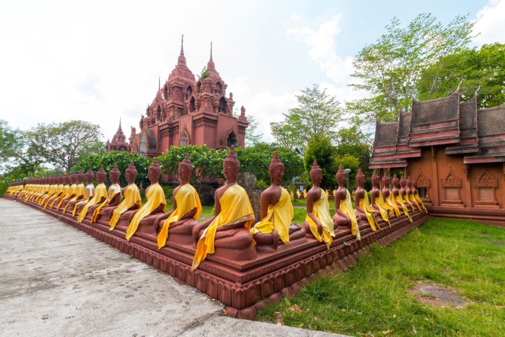 Row of seated golden Buddha statues in yellow robes lining the path to the red-brick temple towers at Wat Khao Angkhan, Buriram